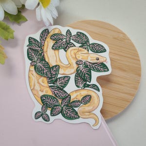 Może przedstawiać: Naklejka przedstawiająca żółtego węża oplecionego zielonymi liśćmi z różowymi żyłkami. Wąż ma szczegółowy wzór i różowy język. Naklejka znajduje się na białym tle z drewnianą powierzchnią i różową powierzchnią.
