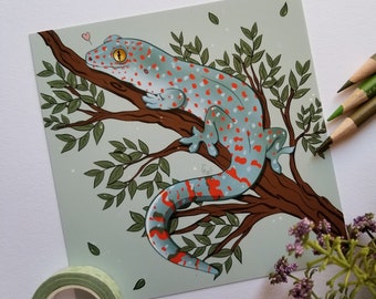 May include: A blue and red gecko with orange eyes perched on a branch of a green leafy tree. The gecko has red and orange spots on its body. The background is a light blue color.