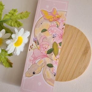 May include: A pink and white bookmark featuring a snake wrapped around pink flowers and a yellow butterfly. The bookmark has a pink background with white stars.
