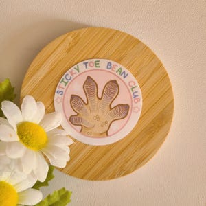 Pode incluir: Um adesivo rosa e branco com uma impress&atilde;o de p&eacute; de gecko de desenho animado e o texto "STICKY TOE BEAN CLUB".