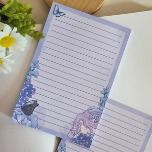 May include: Purple lined notepad with a unicorn, butterflies, and forget-me-not flowers on the cover.
