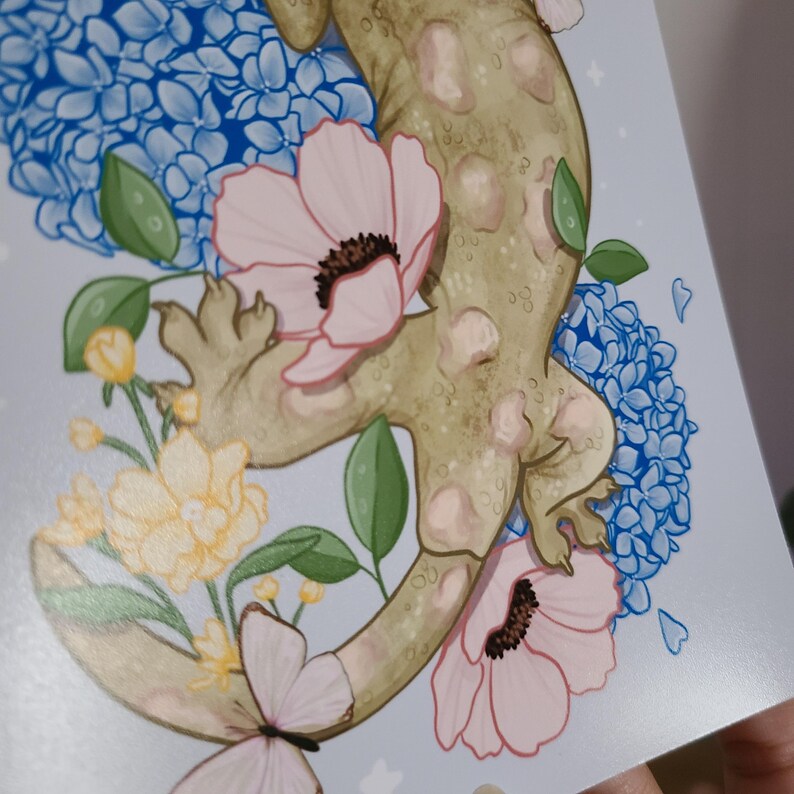 Op de afbeelding: Een artistieke print met een gestileerde gekko verweven met blauwe hortensia's, roze anemonen en gele bloemen. De gekko is lichtgroen met donkere vlekken. De achtergrond is lichtblauw met kleine witte sterren.
