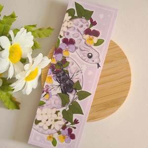 May include: A rectangular bookmark featuring a white snake with purple spots, surrounded by flowers and a black beetle on a light purple background. The bookmark has a whimsical, illustrated style, with a cute snake face and floral accents.