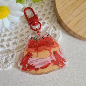 May include: A red, white, and pink strawberry shortcake charm with three red frogs on top. The charm is attached to a red metal key chain.
