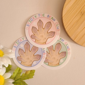 Könnte beinhalten: Drei runde Aufkleber mit einem Cartoon-Gecko-Fußabdruck in der Mitte. Die Aufkleber sind in Pink, Blau und Grün mit dem Text "Sticky Toe Bean Club" am Rand.