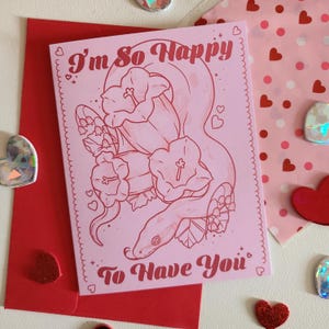 May include: A pink greeting card with the text "I'm So Happy To Have You" in red script. The card features a line drawing of a snake intertwined with flowers. The card is placed on a red envelope, with heart-shaped decorations.
