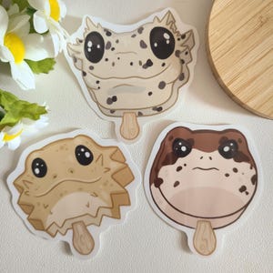 May include: Three different animal popsicle stickers. The first sticker is a white gecko with black spots, the second is a brown frog with black spots, and the third is a light brown bearded dragon with black eyes.
