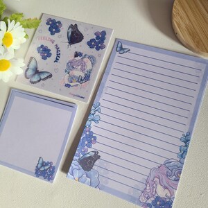 May include: A stationery set featuring a purple and blue colour scheme with a unicorn, butterflies, and forget-me-not flowers. The set includes a sheet of stickers with the text "Feeling Magical", a notecard, and a sheet of lined paper.