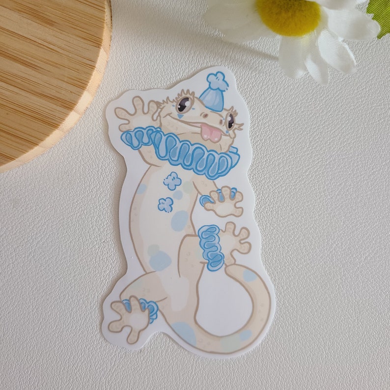 May include: A white gecko wearing a blue and white jester hat and a blue ruffled collar. The gecko has blue spots and is standing on its hind legs with its front paws raised.