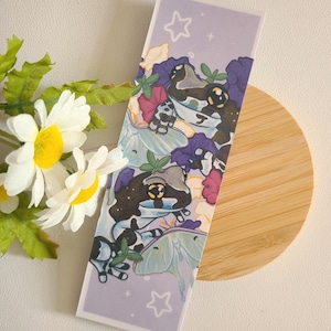 May include: A purple and blue bookmark with two white and black frogs, a moth, and stars. The bookmark is on a light brown wooden surface.