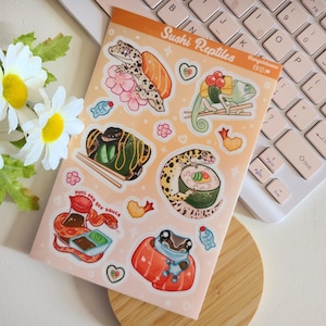 May include: A sheet of stickers featuring various reptiles dressed as sushi. The stickers are on a pink background with white hearts and flowers. The text "Sushi Reptiles" is at the top of the sheet.