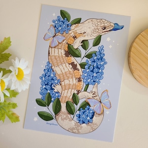 Może przedstawiać: Cyfrowa ilustracja jaszczurki z niebieskim językiem o jasnobrązowym i beżowym ciele w wzory. Jaszczurka otoczona jest niebieskimi i fioletowymi kwiatami oraz motylami. Tło jest jasnoniebieskie.