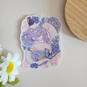 May include: A white snake with purple and pink accents is curled around a bouquet of purple flowers and butterflies. The snake has a purple and pink head with a pointed snout. The flowers are surrounded by pink and white petals.