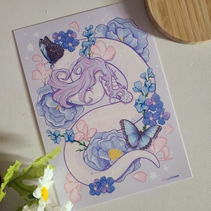 Może przedstawiać: Fioletowy i różowy smok zwinięty w łóżku z niebieskich i białych kwiatów. Smok ma róg i długi ogon. Wokół smoka latają motyle.