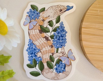May include: A sticker featuring a beige and brown lizard with a blue tongue, surrounded by blue flowers and butterflies. The lizard is climbing up a branch of green leaves.