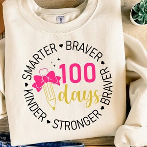 Op de afbeelding: Een wit sweatshirt met een zwart cirkelontwerp met de tekst "Smarter Kinder Braver 100 days Stronger Braver". Binnen de cirkel bevindt zich een roze strik met een gouden potlood.