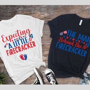 May include: Two white and navy blue t-shirts with red, white, and blue text and graphics. The white t-shirt says "Expecting a little firecracker" with a red, white, and blue firework graphic and a pair of baby footprints. The navy blue t-shirt says "The man behind the firecracker" with a red, white, and blue firework graphic and sunglasses.