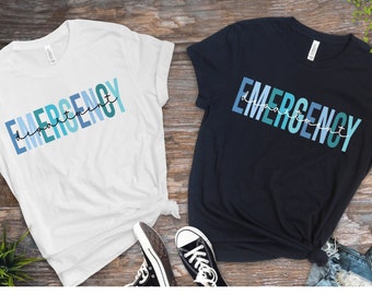 May include: Two t-shirts with the text "Emergency Department" in a gradient of blue colors. The shirts are white and navy blue.