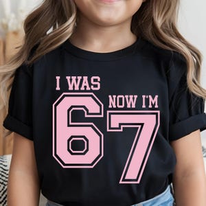 Könnte beinhalten: Schwarzes T-Shirt mit dem Text "I WAS 67 NOW I'M 67" in Rosa. Die Zahl 67 ist in einer fetten Schriftart im College-Stil gehalten. Das Shirt ist wahrscheinlich für eine Geburtstagsfeier.