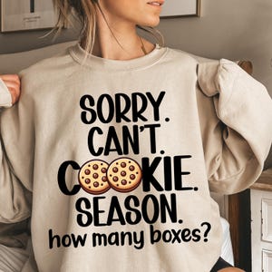Könnte beinhalten: Beigefarbener Pullover mit dem Text "Sorry. Can't. Cookie. Season. How many boxes?" und zwei Schokoladenkeksen.