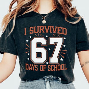 May include: Dark gray t-shirt with the text "I SURVIVED 67 DAYS OF SCHOOL" in orange and white. The number 67 is in a bold, outlined font, with star accents. The shirt is a casual, crew neck style.