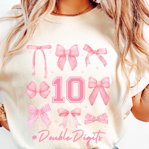 May include: A white t-shirt with a pink number 10 and pink bows. The text "Double Digits" is printed below the number.