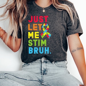 May include: A dark grey t-shirt with the text "JUST LET ME STIM BRUH." in colorful letters. A rainbow puzzle piece ribbon is next to the text.