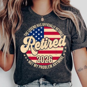 Może przedstawiać: Ciemnoszara koszulka z okrągłą grafiką przedstawiającą amerykańską flagę. Grafika zawiera słowo "Retired" w stylu vintage, rok "2026" i frazę "Not My Problem Anymore". Koszulka ma okrągły dekolt.