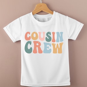 Könnte beinhalten: Weißes T-Shirt mit dem Schriftzug "COUSIN CREW" in bunten, Retro-Buchstaben. Der Text ist in Pfirsich-, Gelb-, Rosa-, Türkis- und Blautönen gehalten. Das Shirt hängt an einem Holzkleiderbügel.