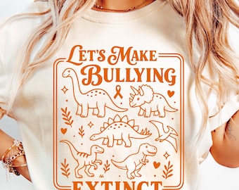 Puede incluir: Una camiseta de color crema con un estampado gráfico en naranja quemado. El diseño incluye el texto "LET'S MAKE BULLYING EXTINCT" rodeado de ilustraciones de cinco dinosaurios diferentes, corazones pequeños, estrellas y ramitas.