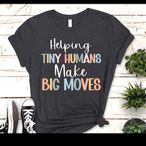 Puede incluir: Una camiseta gris oscuro jaspeada con el texto "Helping Tiny Humans Make Big Moves" en letras de colores.