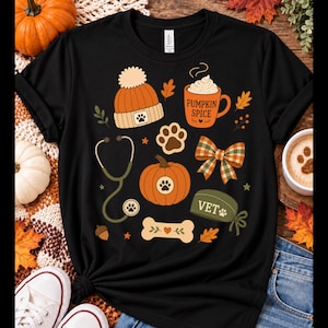 Puede incluir: Una camiseta negra con un estampado gráfico de artículos veterinarios con temática otoñal que incluye un gorro naranja, una taza con la etiqueta PUMPKIN SPICE, un estetoscopio, una calabaza con una huella, un lazo a cuadros, un hueso y una gorra verde con VET.