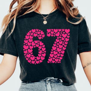 May include: Black t-shirt with the number "67" in bright pink hearts. The shirt has a crew neck and short sleeves. The model is wearing a gold necklace and blue jeans.