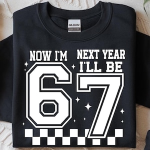 May include: Black sweatshirt with white text that reads "NOW I'M 66 NEXT YEAR I'LL BE 67". The numbers are large and outlined in white, with a checkered pattern at the bottom. The sweatshirt is a Gildan Heavy Cotton.