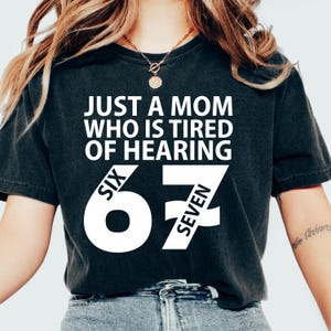 Könnte beinhalten: Dunkelgraues T-Shirt mit dem weißen Text "JUST A MOM WHO IS TIRED OF HEARING SIX 67 SEVEN". Das T-Shirt hat einen Rundhalsausschnitt und kurze Ärmel. Der Text ist fett und ohne Serifen.