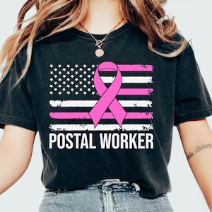 May include: Black t-shirt featuring a distressed American flag design with a pink ribbon and the text "POSTAL WORKER" in white. The shirt has a crew neck and short sleeves.