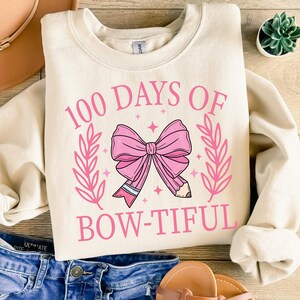May include: Cream-colored sweatshirt with the text "100 Days of Bow-tiful" in pink. The design features a pink bow and pencil graphic, surrounded by pink leaves and stars. The sweatshirt is folded on top of blue jeans.