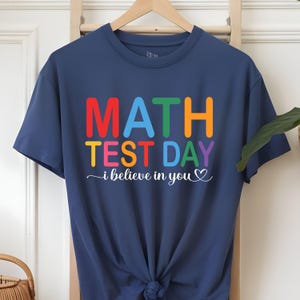 Puede incluir: Camiseta azul marino con las palabras "MATH TEST DAY" en letras de colores. Debajo, la frase "i believe in you" está escrita en cursiva con un corazón. La camiseta está colgada en una percha de madera.