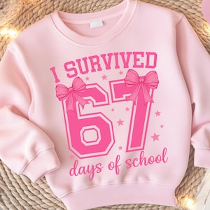 May include: A light pink sweatshirt with the text "I SURVIVED 67 days of school" in a bright pink font. The number "67" is decorated with bows and stars. The sweatshirt has long sleeves and a crew neck.