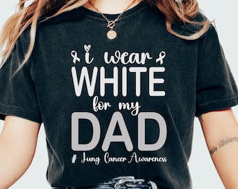 Puede incluir: Camiseta gris oscuro con el texto "I wear WHITE for my DAD" en blanco y gris. La camiseta también incluye el texto "# Jung Cancer Awareness". La camiseta es una prenda informal.