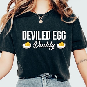 Puede incluir: Camiseta gris oscuro con la frase "DEVILED EGG Daddy" en blanco. El diseño incluye dos ilustraciones de huevos rellenos. Es una camiseta de estilo casual y cómodo.