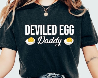 Pode incluir: T-shirt cinza escuro com a frase "DEVILED EGG Daddy" em branco. O design inclui duas ilustrações de ovos recheados. É uma camiseta de estilo casual e confortável.