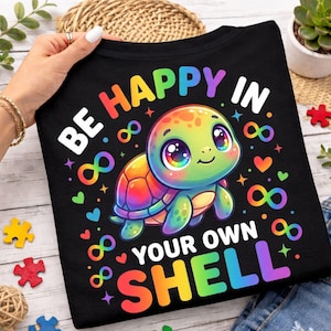 May include: Black t-shirt featuring a vibrant cartoon turtle with a rainbow shell and the text "BE HAPPY IN YOUR OWN SHELL". The design includes hearts and infinity symbols in various colours, creating a cheerful aesthetic.