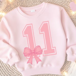 May include: Pink sweatshirt with a large pink number "11" printed on the front. The number has a white outline and a pink bow is centered below the number.