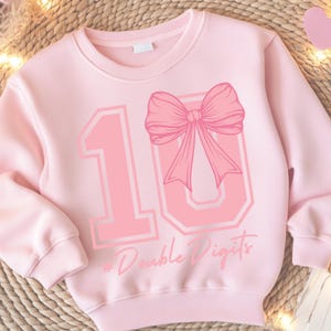 Op de afbeelding: Roze trui met een groot roze nummer 10 met een roze strik erop. De tekst "#Double Digits" staat onder het nummer.