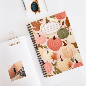 Op de afbeelding: Een roze spiraalnotitieblok met een herfstthema met pompoenen, bladeren en bloemen. Het notitieblok heeft een blanco label aan de voorkant.