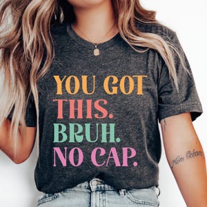 以下が含まれることがあります： 暗いグレーのTシャツに「YOU GOT THIS. BRUH. NO CAP.」という文字がカラフルな文字でプリントされています。文字はオレンジ、ピンク、グリーン、ライトピンクです。