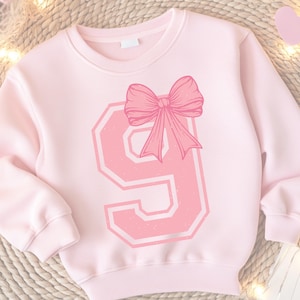 May include: Pink sweatshirt with a large pink number nine and a pink bow on top. The number nine has a white outline and a distressed look.