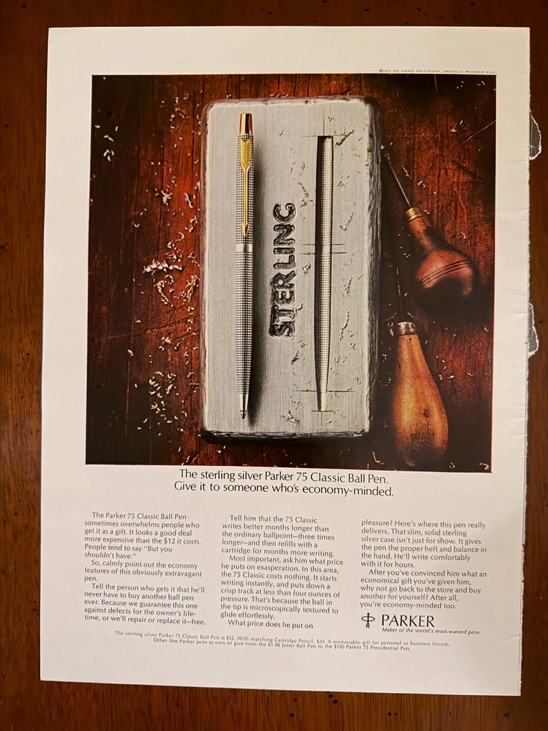 Peut inclure: Publicit&eacute; pour un stylo &agrave; bille Parker 75 Classic. Le stylo argent&eacute; avec des d&eacute;tails dor&eacute;s est pr&eacute;sent&eacute; sur un &eacute;tui argent&eacute; avec le mot "STERLING" en relief. Le texte met en avant les caract&eacute;ristiques et la valeur du stylo.