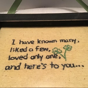 May include: A framed piece of art with a black frame and a beige background. The art features a handwritten quote that reads "I have known many, liked a few, loved only one, and here's to you..." with two green shamrocks embroidered on the right side.